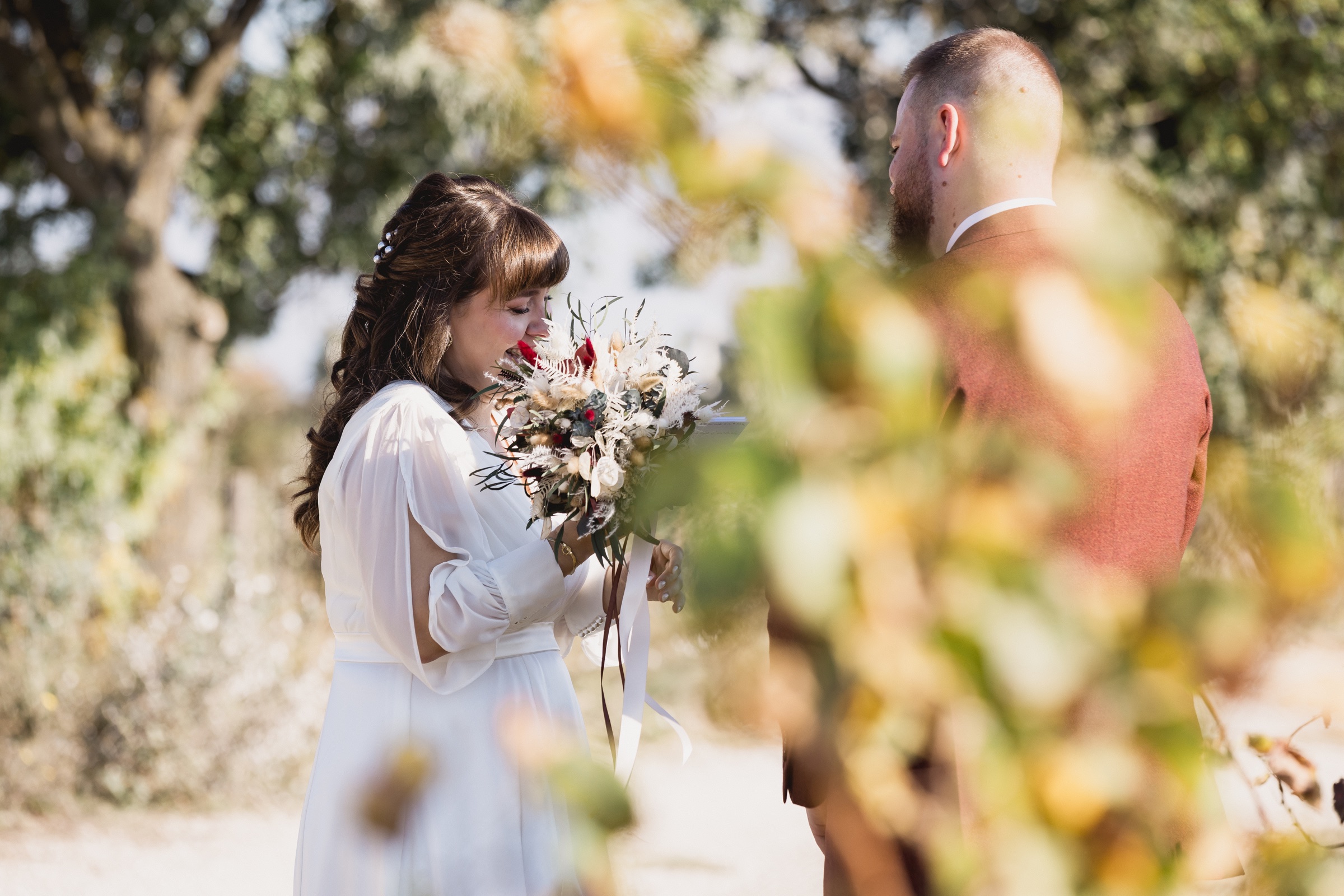 Mariée tenant son bouquet, photographiée à travers des feuillages